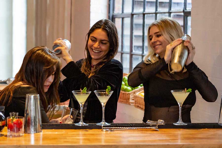 Three women on a hen do stood behind a cocktail bar making cocktails